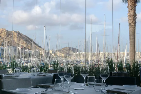 Las mesas de la terraza de Dársena con las copas puestas, y los mástiles del puerto deportivo y la ladera del castillo al fondo